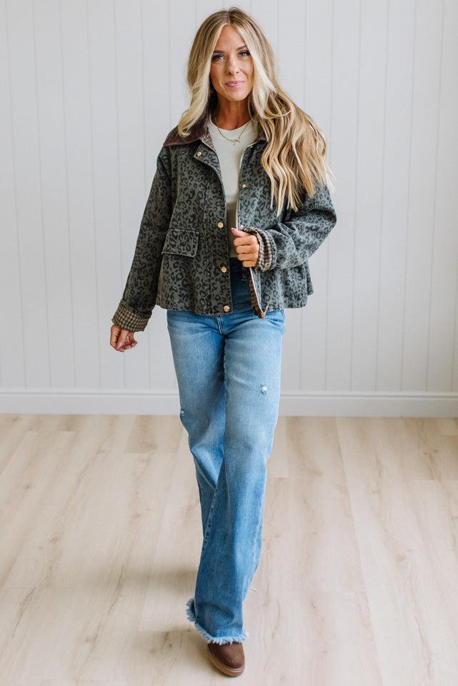 Blonde woman wearing a leopard print barn jacket with brown corduroy collar and gingham plant lining. Standing on a light wood floor with panel backdrop.