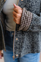 Blonde woman wearing a leopard print barn jacket with brown corduroy collar and gingham plant lining. Standing on a light wood floor with panel backdrop.