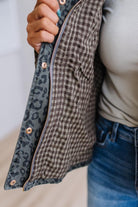 Close-up of a person holding open a quilted jacket, showing a brown and black gingham pattern on the outside and a blue-gray leopard print lining with rose gold snap buttons.