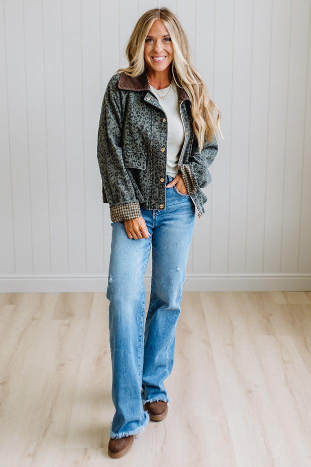 Blonde woman wearing a leopard print barn jacket with brown corduroy collar and gingham plant lining. Standing on a light wood floor with panel backdrop.