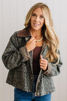 A woman modeling a cropped leopard-print jacket with a brown sherpa collar, worn over a brown top and blue jeans against a white paneled wall.