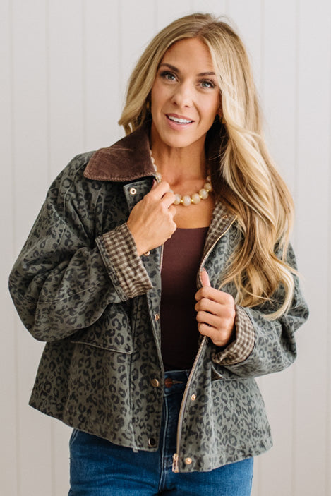A woman modeling a cropped leopard-print jacket with a brown sherpa collar, worn over a brown top and blue jeans against a white paneled wall.