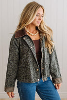 A woman wearing a cropped leopard-print jacket with a brown corduroy collar, styled with a brown top, blue jeans, and a pearl necklace against a white paneled wall.