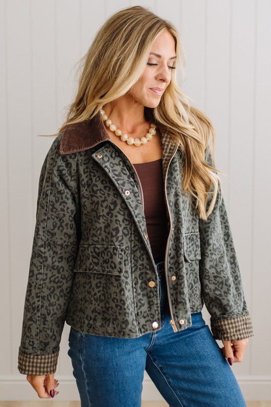 A woman wearing a cropped leopard-print jacket with a brown corduroy collar, styled with a brown top, blue jeans, and a pearl necklace against a white paneled wall.