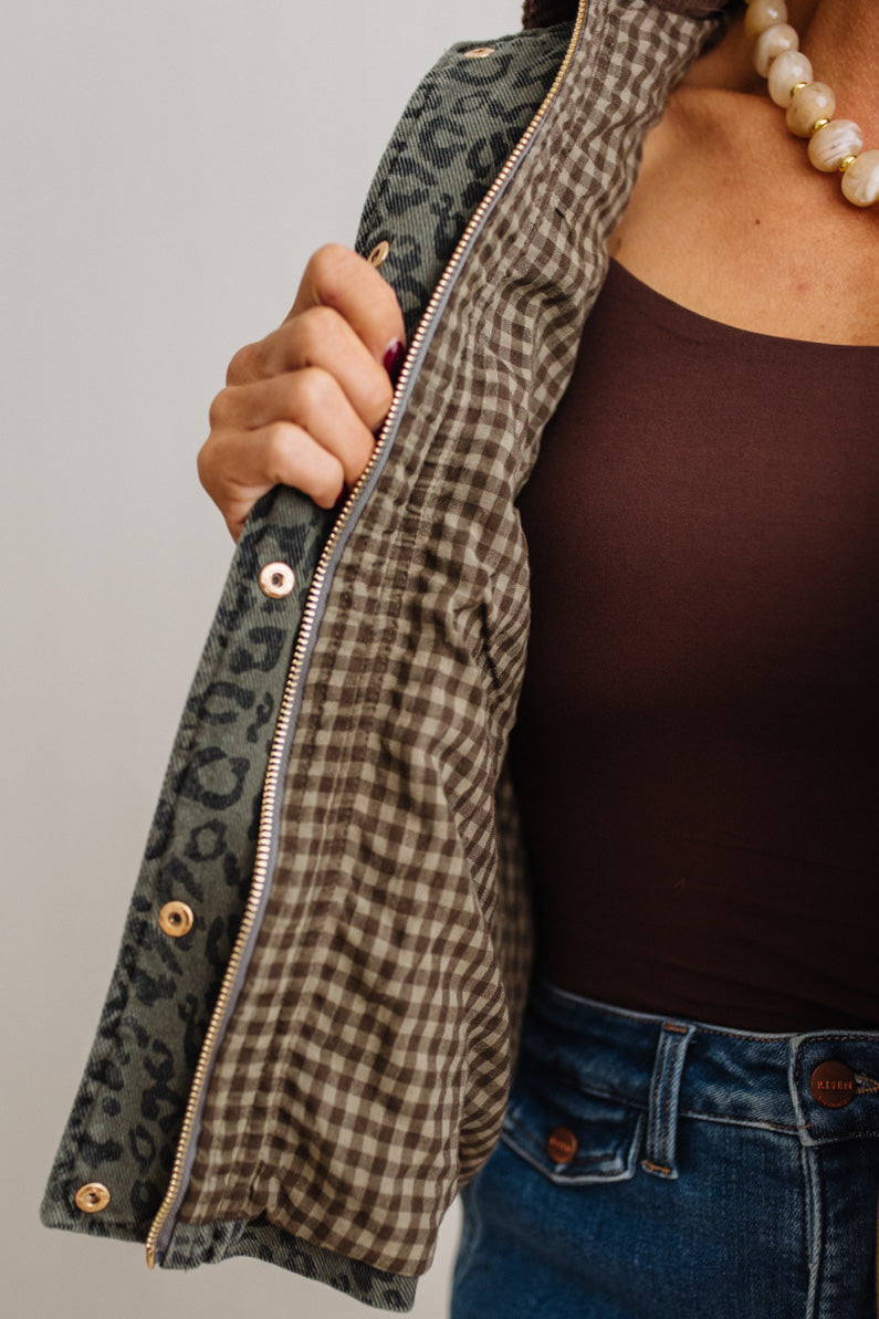 Close-up of a person wearing a zip-front jacket with a leopard-print outer panel and a brown-and-white gingham lining, shown against a plain light gray background.