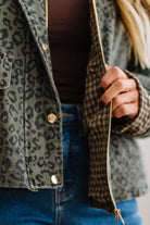Close-up of a person wearing a cropped olive green leopard-print jacket with gold-tone snap buttons and a gold zipper, layered over a dark brown top and blue jeans.