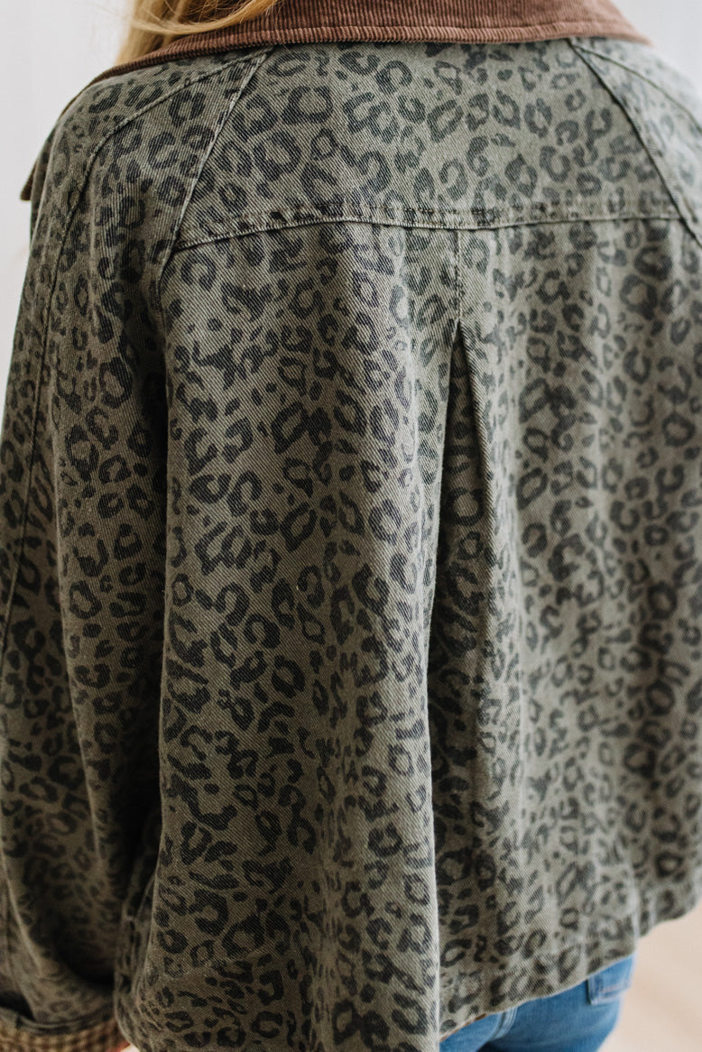 Close-up back view of a person wearing a cropped leopard-print jacket in muted olive green and black, with a brown corduroy collar and blue jeans visible at the bottom.
