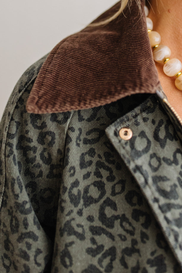 Close-up of a person wearing a corduroy jacket with a brown collar and a muted green leopard print body, paired with a pearl necklace.