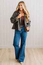 A woman modeling a cropped patterned jacket with a brown collar, worn over a brown top and blue flare jeans, standing against a white paneled wall and light wood floor.