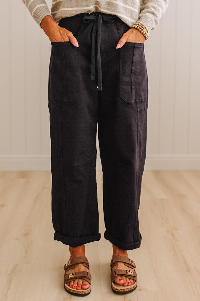 A person wearing dark charcoal wide-leg pants with a drawstring waist, front patch pockets, and rolled cuffs, paired with brown slide sandals in a bright indoor room with light wood flooring.