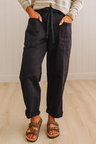 A person wearing dark charcoal cargo-style pants with a drawstring waist, shown from the waist down in a home setting with light wood flooring and paneled walls.