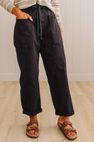 A person wearing loose black wide-leg pants with a drawstring waist, side pockets, and cuffed hems, paired with brown sandals and a light striped top, standing on a light wood floor against a white paneled wall.