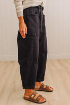 A person wearing loose black drawstring pants with side cargo pockets, paired with a striped long-sleeve top and brown slide sandals, standing on a light wood floor against a white paneled wall.