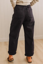 A person shown from the back wearing loose black wide-leg pants with cuffed hems, a cream sweater, and brown wedge sandals in a bright indoor room with light wood flooring and white paneled walls.