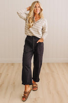 A woman modeling a beige and white striped long-sleeve henley top with dark charcoal wide-leg pants and brown slide sandals, standing in a room with light wood floors and white paneled walls.