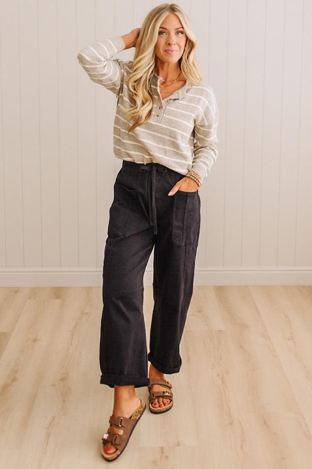 A woman modeling a beige and white striped long-sleeve henley top with dark charcoal wide-leg pants and brown slide sandals, standing in a room with light wood floors and white paneled walls.