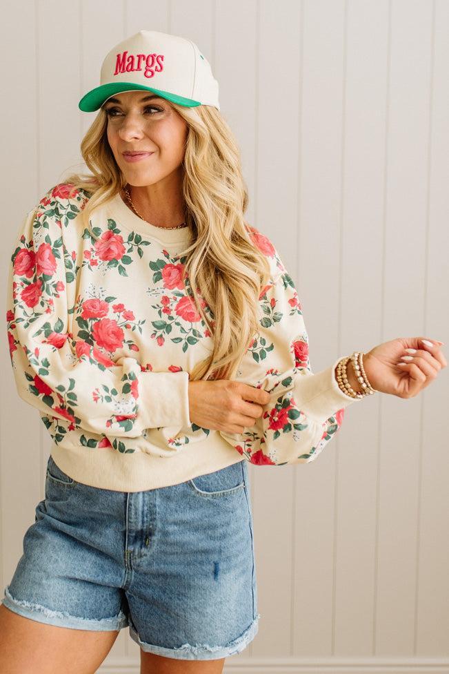 Woman wearing a floral sweatshirt, denim shorts, and a cap with 'Margs' text.