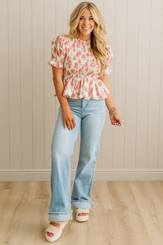 Woman wearing a floral top and light blue jeans against a neutral wall.