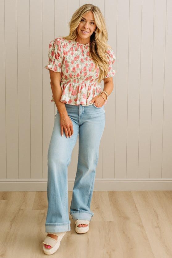Woman wearing a floral top and light blue jeans standing against a beige wall.