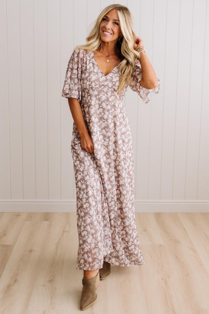 A woman modeling a long-sleeve maxi dress with a dusty rose and white floral pattern, standing in a room with light wood floors and white paneled walls.