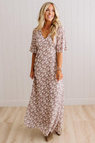 A woman modeling a long, short-sleeve maxi dress with a dusty rose and white floral pattern, standing in front of a white paneled wall and light wood floor.