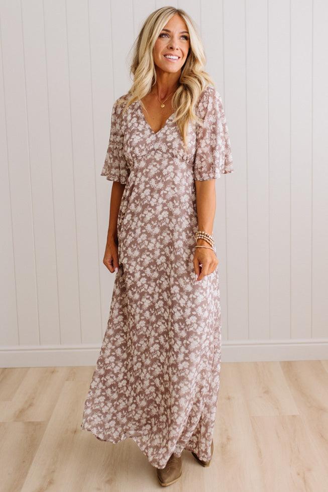 A woman modeling a long, short-sleeve maxi dress with a dusty rose and white floral pattern, standing in front of a white paneled wall and light wood floor.