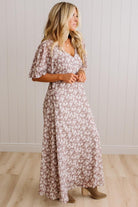 A woman modeling a long, flowy maxi dress with a dusty rose and white floral pattern, standing in front of a white paneled wall and light wood floor.