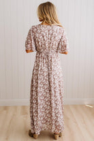 A woman viewed from behind wearing a knee-length floral dress with short puff sleeves and a tie waist, standing on a light wood floor against a white paneled wall.