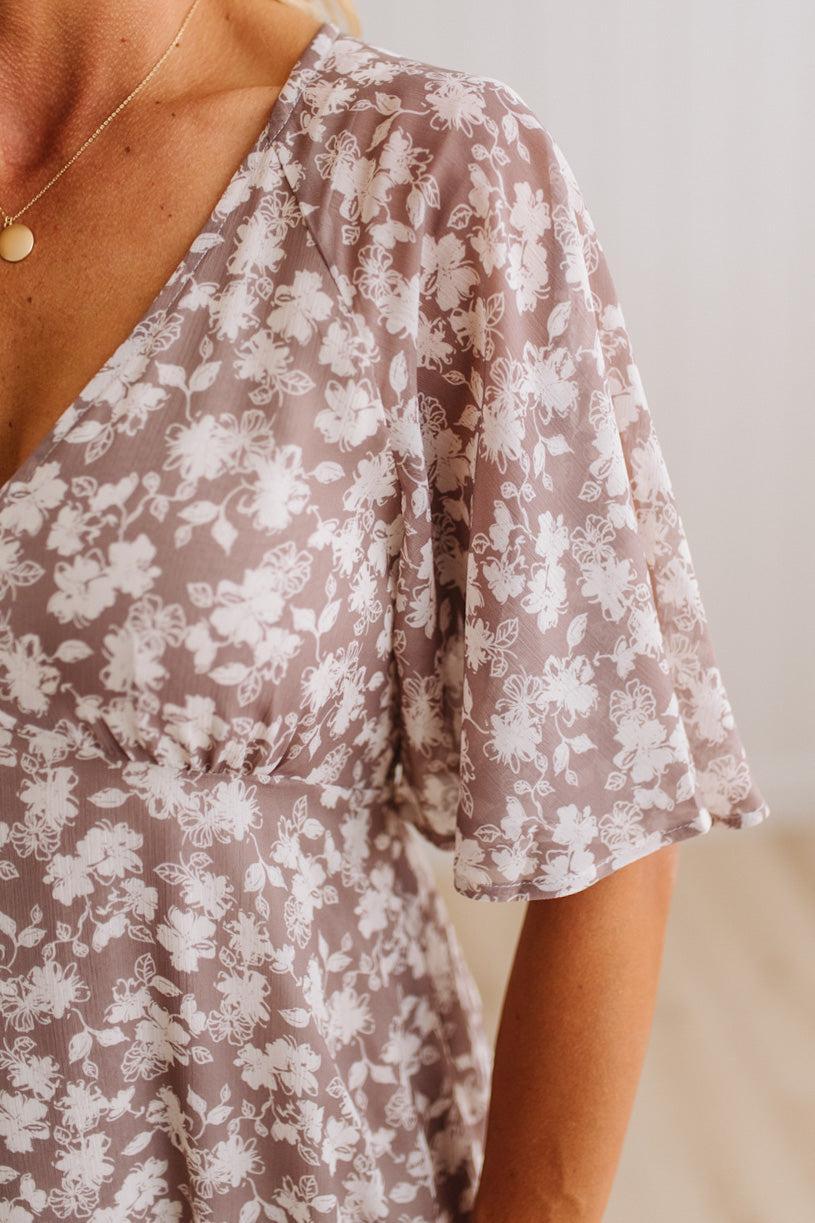 Close-up of a person wearing a sheer taupe-brown V-neck dress with a white floral print and flutter sleeves.