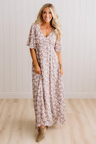 A woman modeling a long-sleeve, V-neck maxi dress with a dusty rose and white floral pattern, standing in front of a white paneled wall and light wood floor.