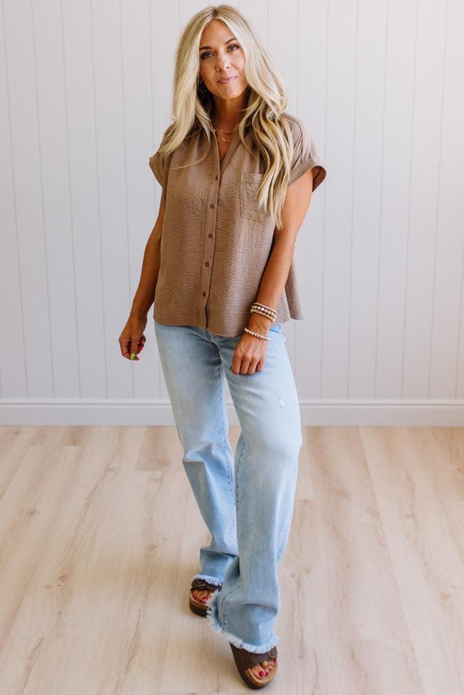 Woman wearing a brown button-up shirt and light blue jeans standing on a wooden floor.