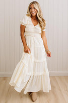 A woman wearing a cream-colored, tiered maxi dress with short puff sleeves and a smocked bodice, standing in front of a white paneled wall and light wood floor.