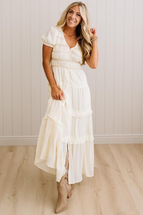 A woman modeling a cream-colored, tiered maxi dress with short puff sleeves and a smocked bodice, paired with beige ankle boots, set against a light wood-paneled wall and floor.