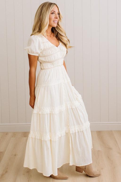 A woman modeling a cream-colored, tiered maxi dress with short puff sleeves and a smocked bodice, standing in front of a white paneled wall and light wood floor.