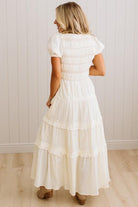 A woman viewed from behind wearing a white tiered maxi dress with short puff sleeves and a gathered waist, standing on a light wood floor against a white paneled wall.