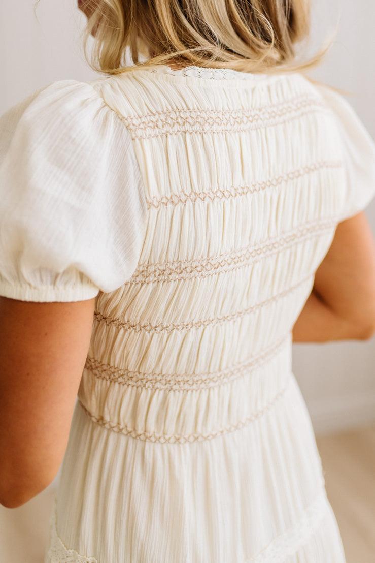 Close-up back view of a woman wearing a cream-colored short-sleeve dress with delicate horizontal smocked and embroidered details across the back.