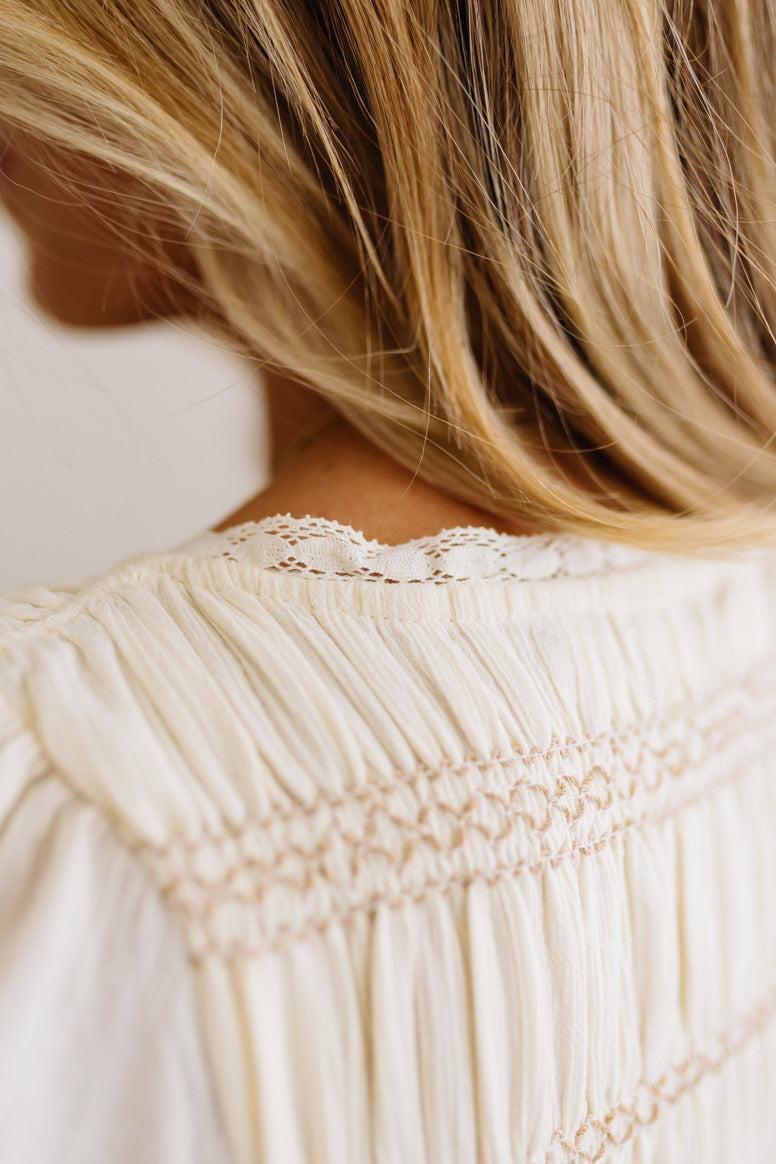 Close-up of a person wearing a cream-colored blouse with delicate lace trim and smocked detailing on the back, with blonde hair visible.