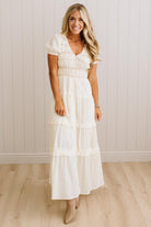 A woman modeling a white tiered maxi dress with short puff sleeves and a smocked bodice, standing in front of a white paneled wall and light wood floor.