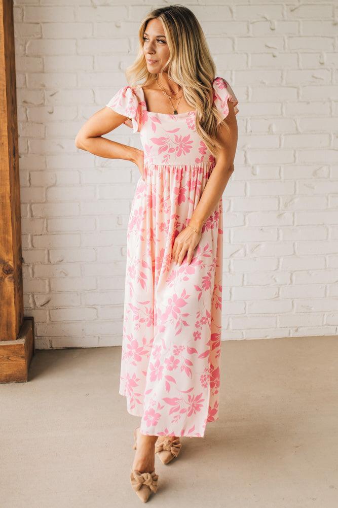 Blonde woman wearing a maxi dress with floral pink and a dark pink print, ruffle sleeved, with back smocked panel.