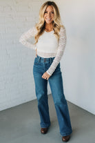 Woman wearing a white lace top and blue jeans against a white brick wall.
