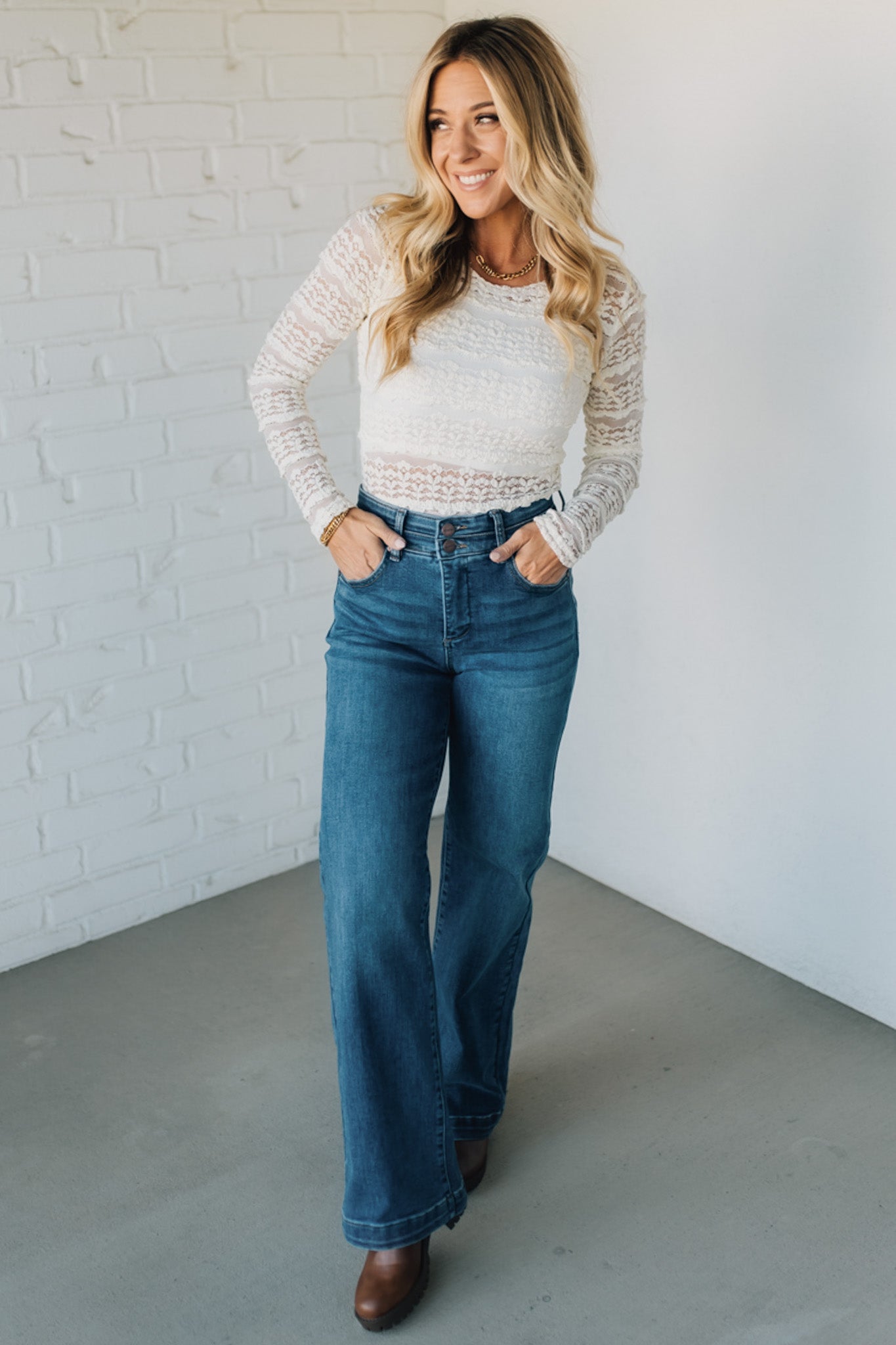 Woman wearing a white lace top and blue jeans standing against a white brick wall.