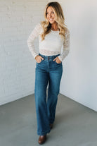 Woman wearing a white lace top and blue jeans standing against a white brick wall.