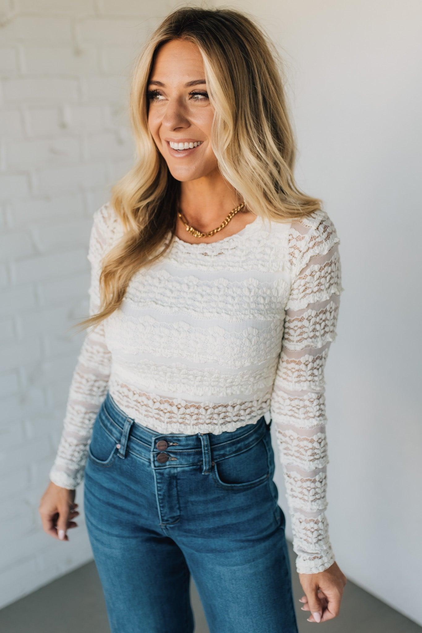 Woman wearing a white lace top and blue jeans against a white wall.