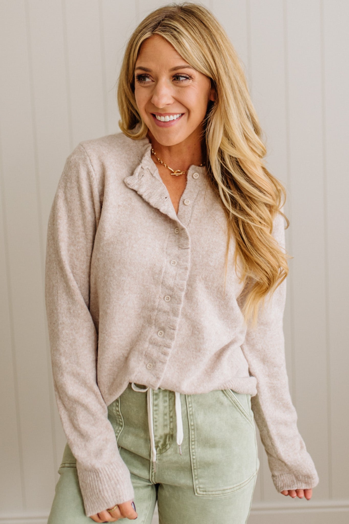 Woman wearing a beige cardigan and green pants against a white wall