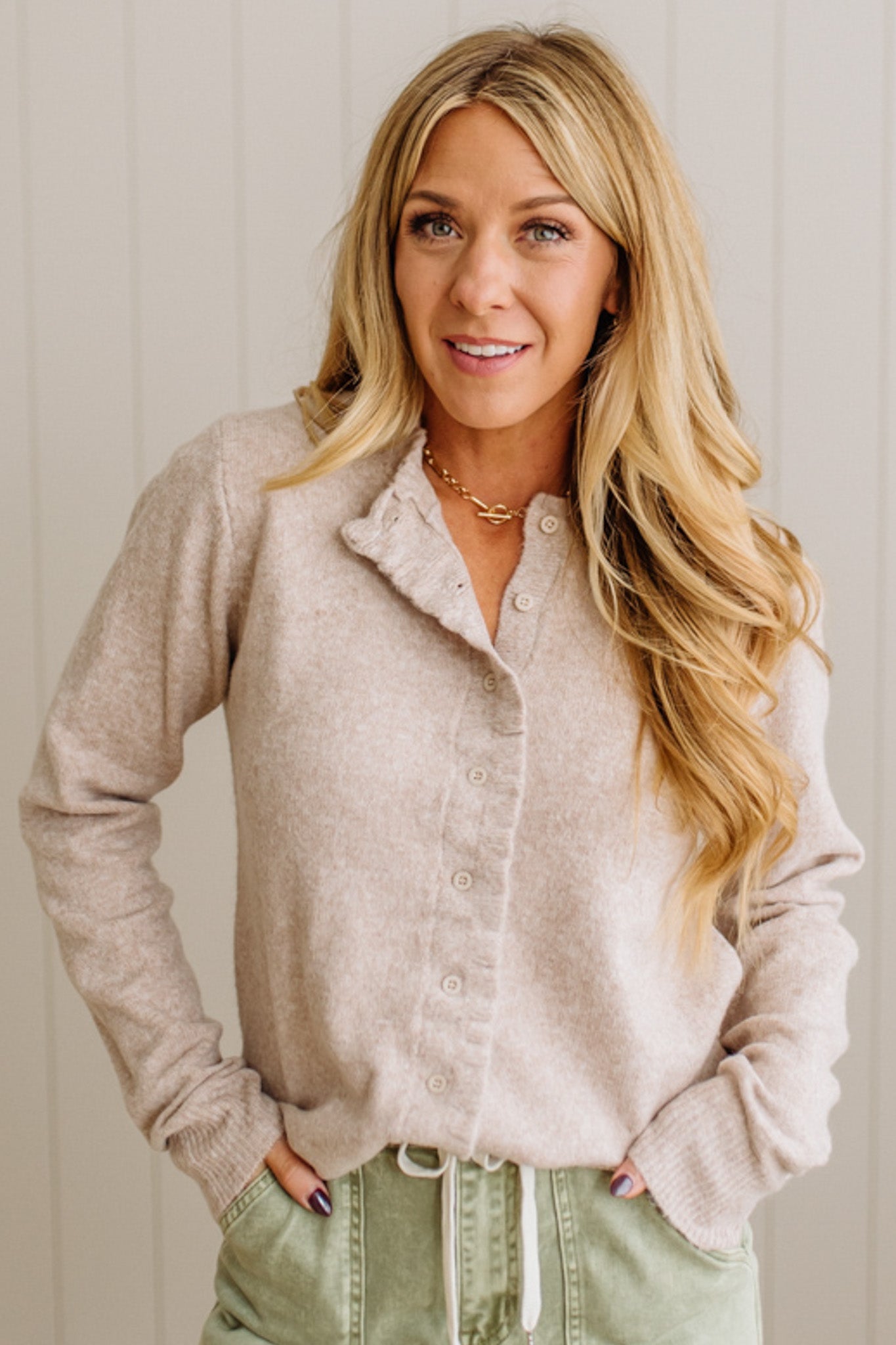 Woman wearing a beige cardigan and green pants against a white wall