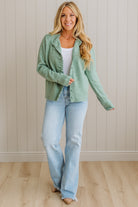 Woman wearing a green jacket and blue jeans against a beige wall.