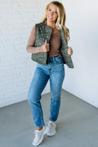 Woman wearing a floral green vest over a brown top and blue jeans against a white brick wall.