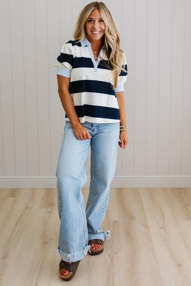 Woman wearing a striped shirt and jeans standing against a white paneled wall.