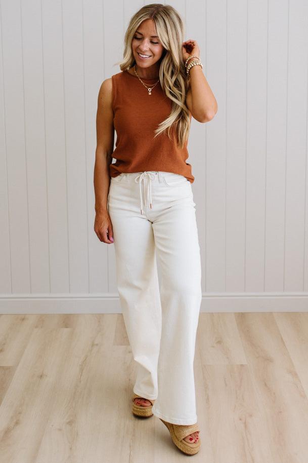 Woman wearing a brown sleeveless top and white pants standing against a light wooden floor and wall.