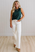 Woman wearing a green sleeveless top and white pants standing against a light wooden paneled wall.